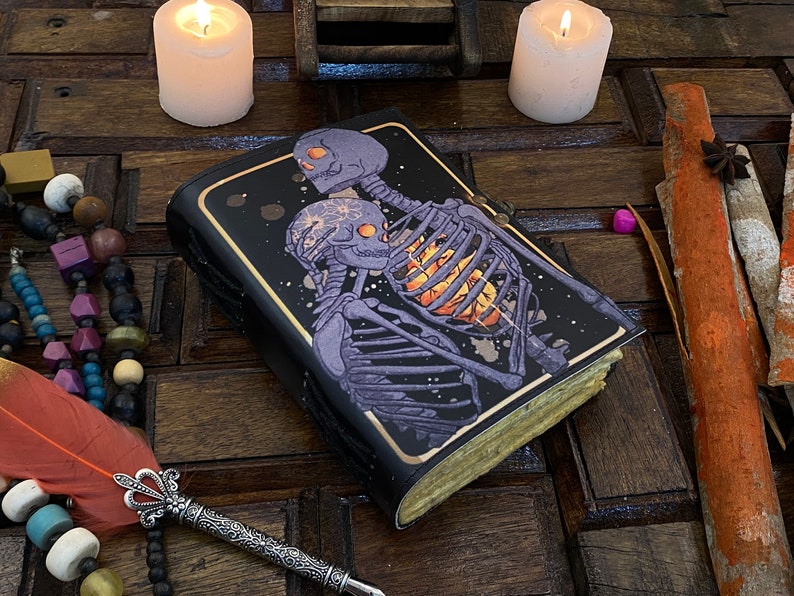 May include: A black leather-bound journal with a dark purple and gold illustration of two skeletons embracing. The skeletons are on a black background with a starry pattern. The journal is on a wooden surface with candles and other decorative items.
