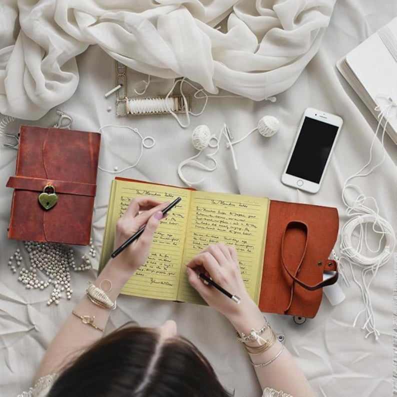 May include: A person writing in a brown leather journal with yellow lined pages. The journal has a lock and a keyhole on the front cover. The journal is surrounded by white yarn, string, and other craft supplies.