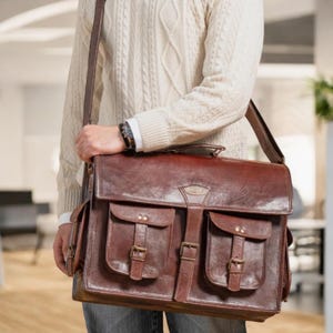May include: A brown leather briefcase with two large front pockets and buckles. The briefcase has a long strap and is worn over the shoulder.