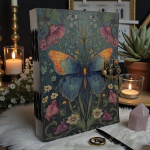 May include: A decorative journal with a butterfly design. The cover features a blue and orange butterfly with floral accents in pink, white, and green. The journal has a metal clasp and is resting on a white fur-like surface.