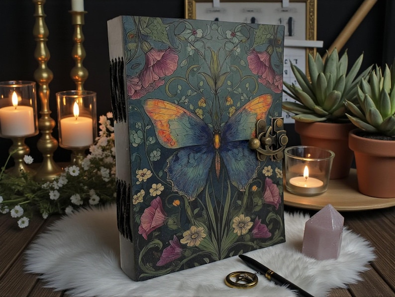 May include: A decorative journal with a butterfly design. The cover features a blue and orange butterfly with floral accents in pink, white, and green. The journal has a metal clasp and is resting on a white fur-like surface.
