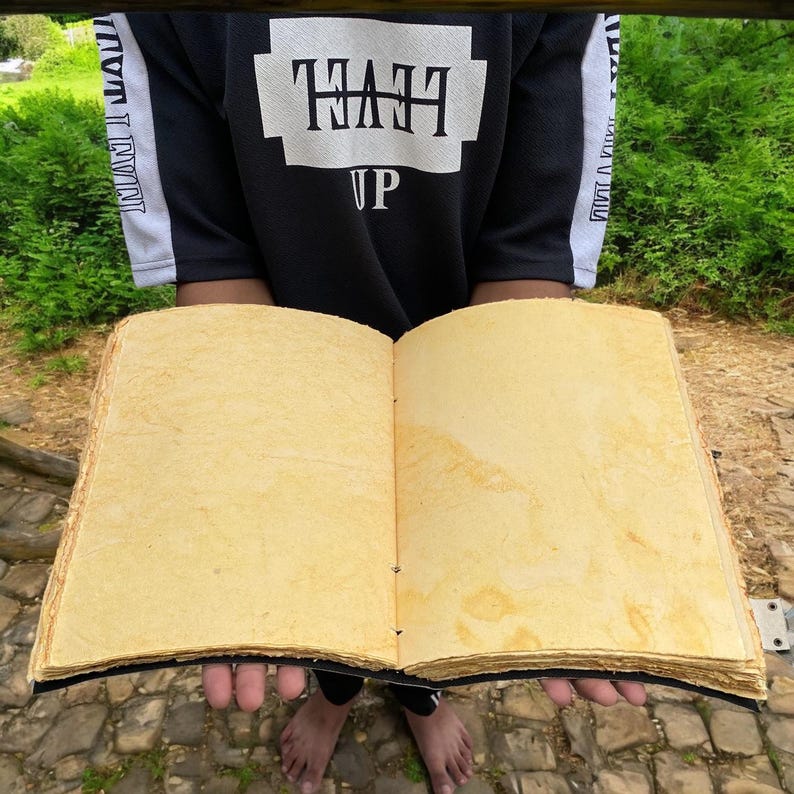May include: A large, open book with thick, yellowed pages. The book is bound in black leather and has a rustic, handmade appearance.