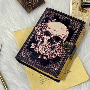 K&ouml;nnte beinhalten: Ein schwarzes Lederjournal mit einem Totenkopf- und Rosenmotiv. Das Journal hat einen Metallverschluss und ist geschlossen.