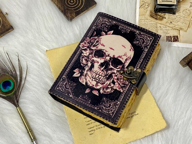 K&ouml;nnte beinhalten: Ein schwarzes Lederjournal mit einem Totenkopf- und Rosenmotiv. Das Journal hat einen Metallverschluss und ist geschlossen.