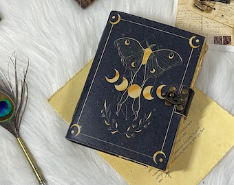 May include: A dark blue journal with a moth and moon phase design in gold. The journal has a metal clasp and is on a white, fluffy surface. A peacock feather and pen are in the background.