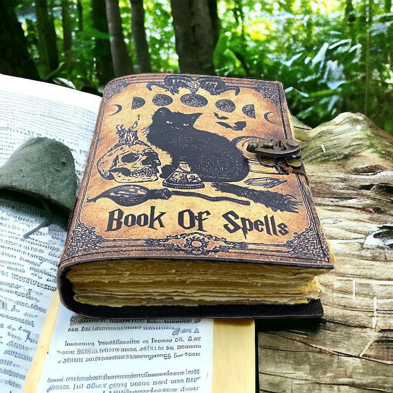 Puede incluir: Un diario encuadernado en cuero marr&oacute;n con un gato negro, una calavera y un dise&ntilde;o de poci&oacute;n. La portada dice "Book of Spells".