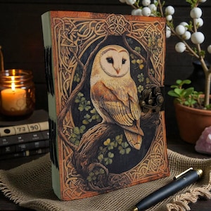 May include: A leather journal with an owl design. The cover shows a barn owl on a branch, framed by Celtic knot patterns and green leaves. It has a metal clasp and a sewn spine. The journal is approximately 20 cm in height.