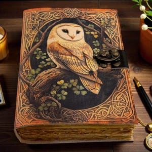 May include: A detailed leather journal with an owl design. The cover depicts a barn owl on a branch, with Celtic knotwork and shamrocks. The journal is secured with a black leather strap. A lit candle, a pen, and a compass are also visible.