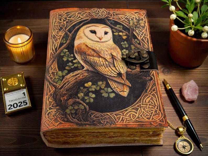 May include: A detailed leather journal with an owl design. The cover depicts a barn owl on a branch, with Celtic knotwork and shamrocks. The journal is secured with a black leather strap. A lit candle, a pen, and a compass are also visible.