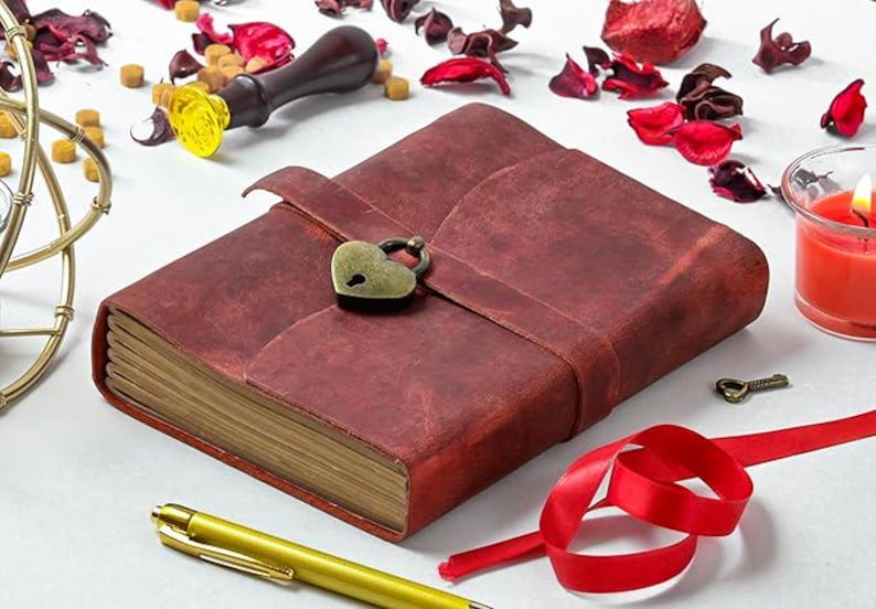 May include: A red leather-bound journal with a heart-shaped lock and key. The journal is closed and has a rustic look. It is surrounded by decorative items, including a wooden stamp, red rose petals, and a candle.