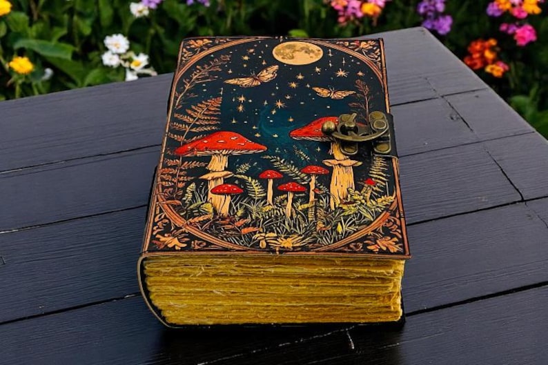 K&ouml;nnte beinhalten: Ein ledergebundenes Notizbuch mit einem skurrilen Pilz- und Schmetterlingsdesign. Das Cover zeigt eine Nachthimmel-Szene mit Vollmond, Sternen und rotkappigen Pilzen. Die Seiten sind gealtert mit einem gelben Farbton, und das Notizbuch wird mit einem Metallverschluss gesichert.