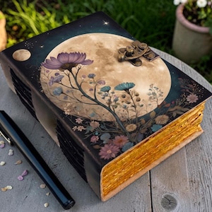 Könnte beinhalten: Ein dekoratives Notizbuch mit einem himmlischen Design mit Vollmond, Blumen und Sternen. Das Notizbuch hat einen dunklen Einband mit einem Messingverschluss und goldfarbenen Kanten. Ein schwarzer Stift liegt neben dem Notizbuch auf einer Holzoberfläche.