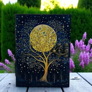 May include: A black leather journal with a gold and blue tree design and a full moon. The moon is yellow and has a tree growing inside of it. The journal has a metal clasp.