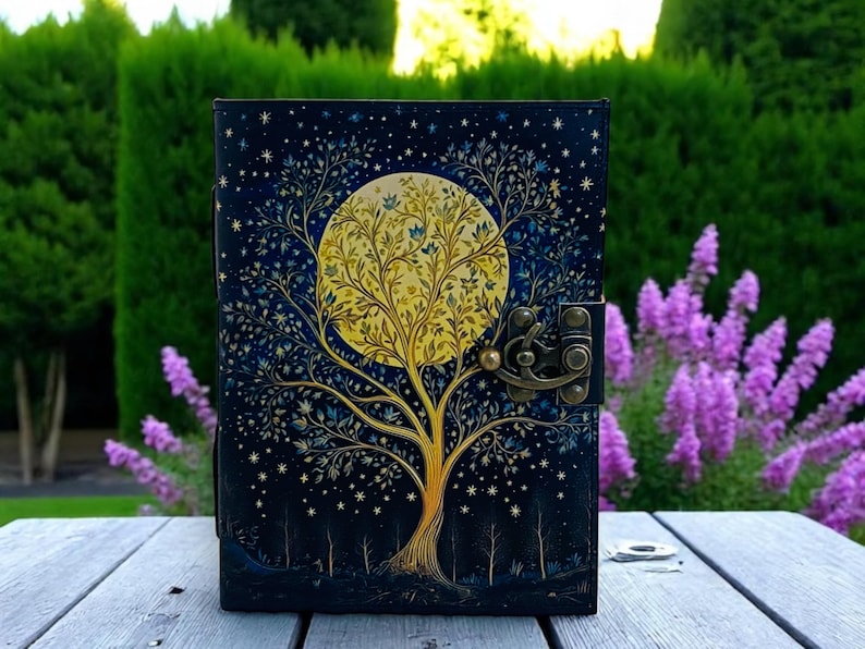 May include: A black leather journal with a gold and blue tree design and a full moon. The moon is yellow and has a tree growing inside of it. The journal has a metal clasp.