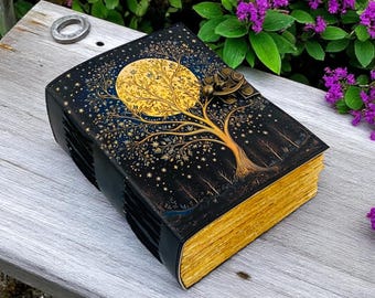 May include: A black leather-bound journal with a gold and blue illustration of a tree with a full moon behind it. The journal has a brass clasp and is closed.