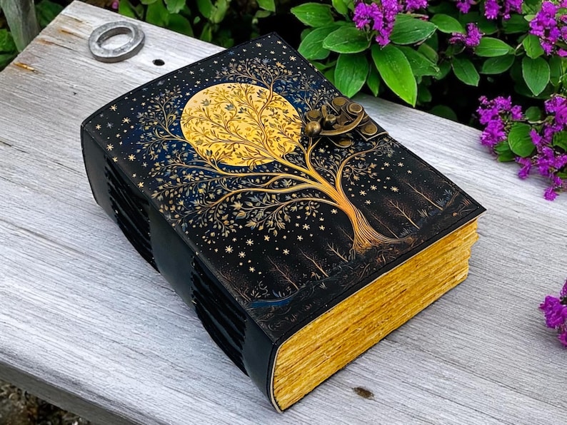 May include: A black leather-bound journal with a gold and blue illustration of a tree with a full moon behind it. The journal has a brass clasp and is closed.