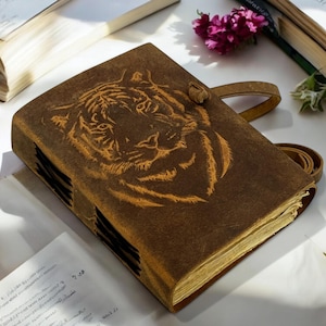 Puede incluir: Un diario de cuero marrón con un diseño de cara de tigre. El diario tiene un cierre de correa y un lazo para un bolígrafo. Las páginas están envejecidas y el diario está rodeado de libros abiertos y flores.