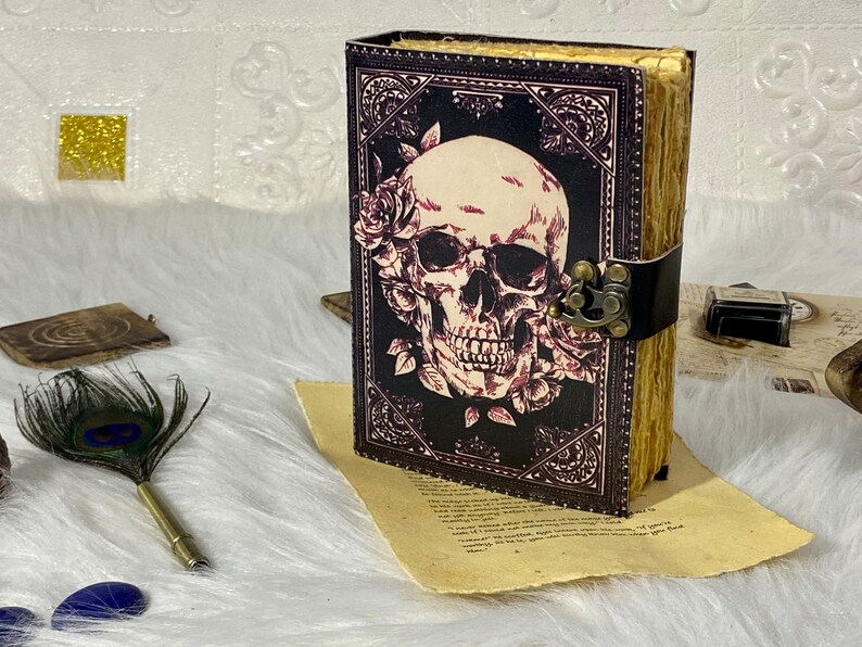 K&ouml;nnte beinhalten: Ein schwarzes, ledergebundenes Tagebuch mit einem Totenkopf- und Rosenmotiv. Das Tagebuch hat einen Metallverschluss und einen dekorativen Rand.