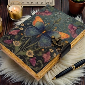 Puede incluir: Un diario decorativo con un diseño de mariposa en la portada, con alas azules y naranjas. El diario está asegurado con una correa y un cierre de cuero negro. Un bolígrafo negro con detalles dorados descansa junto al diario.