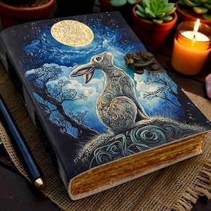 May include: A leather-bound journal with a fantasy illustration of a rabbit under a full moon. The cover features a brass clasp and gold-edged pages. A pen and small dish with white spheres are next to the journal.