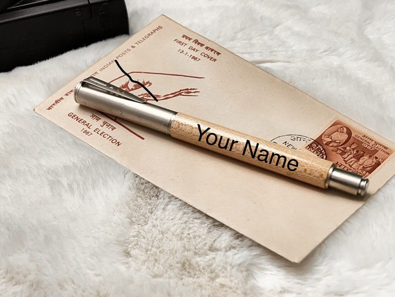 May include: A wooden pen with a silver metal tip and a personalized inscription that reads "Your Name". The pen is resting on a vintage envelope with a stamp featuring a scene of people in traditional Indian clothing.