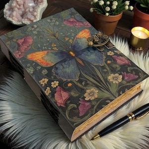 May include: A decorative journal with a butterfly design on the cover. The journal features a brass clasp and is surrounded by flowers in shades of pink, yellow, and green. A black pen rests beside the journal on a white fur surface.