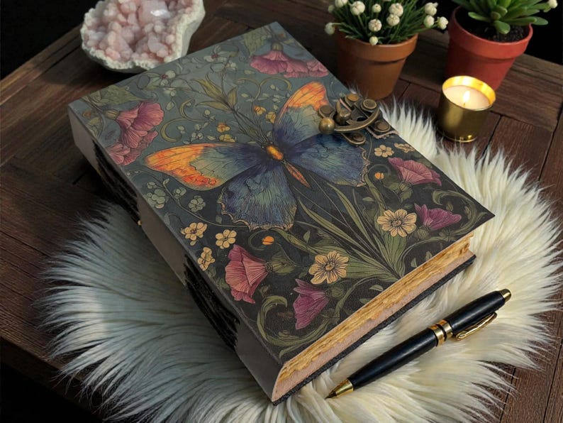 May include: A decorative journal with a butterfly design on the cover. The journal features a brass clasp and is surrounded by flowers in shades of pink, yellow, and green. A black pen rests beside the journal on a white fur surface.