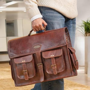 May include: A brown leather briefcase with two large front pockets and a smaller pocket on the side. The briefcase has metal buckles and a top handle.