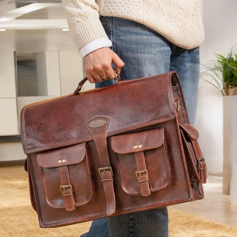 May include: A brown leather briefcase with two large front pockets and a smaller pocket on the side. The briefcase has metal buckles and a top handle.