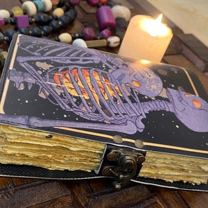 May include: A black leather-bound journal with a skeleton design on the cover. The skeleton is purple and has a glowing orange heart. The journal has a brass clasp and is closed.