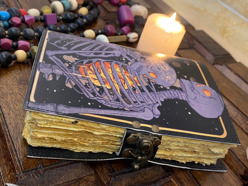 May include: A black leather-bound journal with a skeleton design on the cover. The skeleton is purple and has a glowing orange heart. The journal has a brass clasp and is closed.