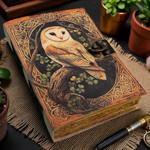 May include: A hardback journal with an owl design. The cover has a detailed Celtic knotwork pattern and a metal clasp. The owl is depicted on a tree branch, surrounded by green clovers. The journal is open, showing aged paper. The journal is surrounded by plants.