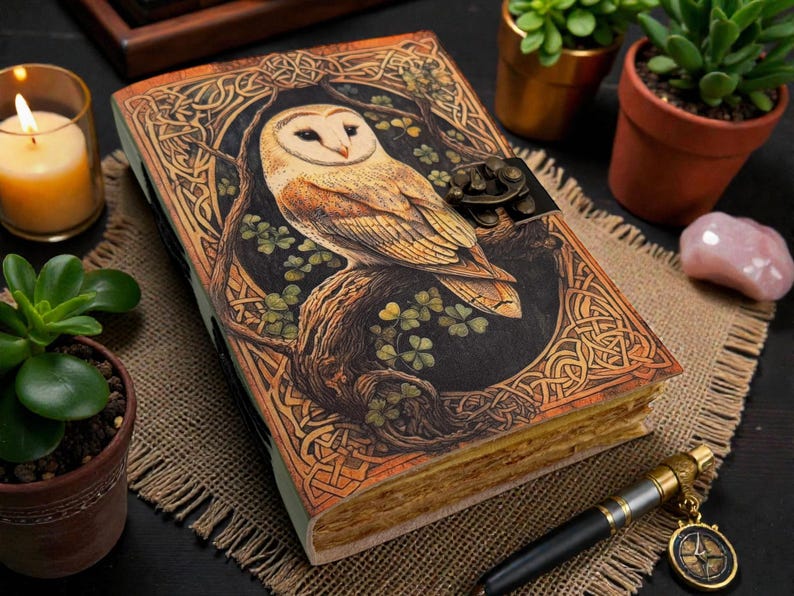 May include: A hardback journal with an owl design. The cover has a detailed Celtic knotwork pattern and a metal clasp. The owl is depicted on a tree branch, surrounded by green clovers. The journal is open, showing aged paper. The journal is surrounded by plants.