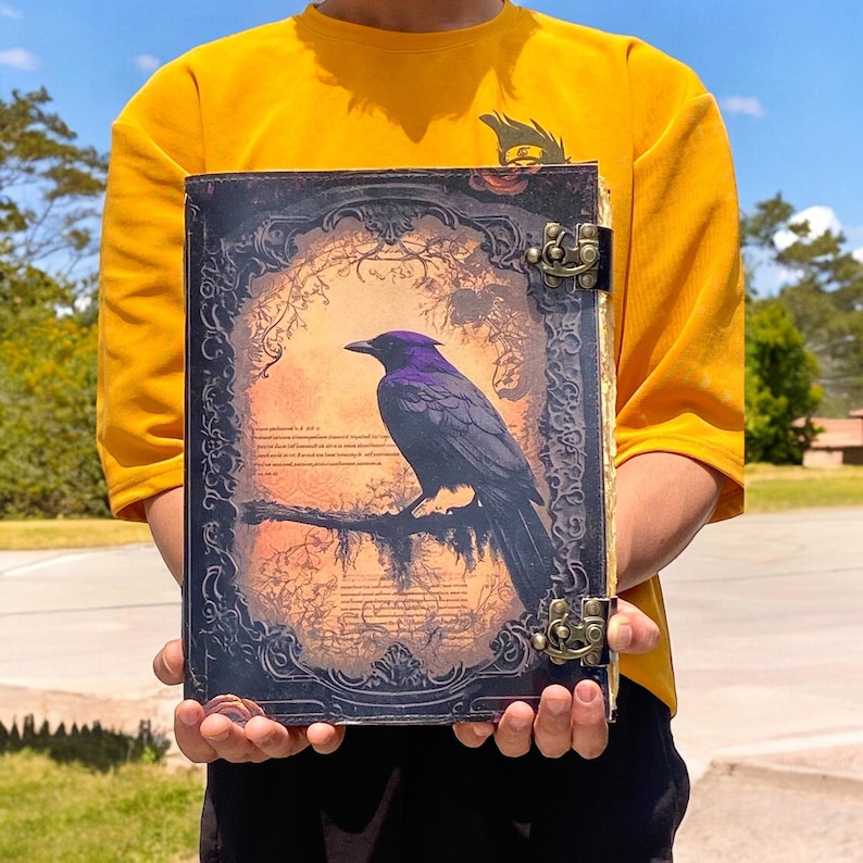 May include: A black leather journal with an ornate design featuring a purple raven perched on a branch. The journal has two brass clasps.