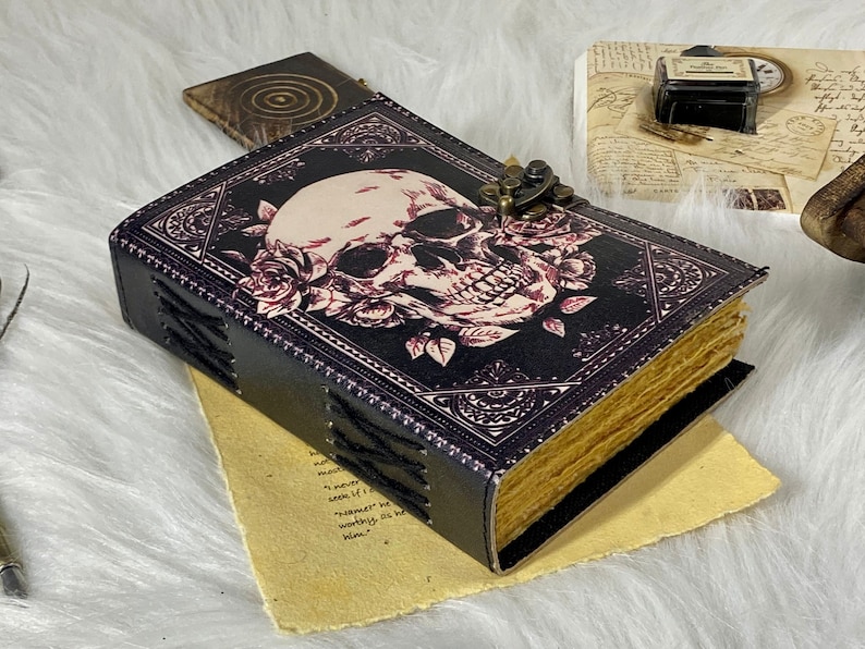 K&ouml;nnte beinhalten: Ein schwarzes, ledergebundenes Tagebuch mit einem Totenkopf- und Blumendesign. Das Tagebuch hat einen Messingverschluss und ist mit schwarzen N&auml;hten verziert.