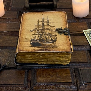 Pode incluir: Um di&aacute;rio de estilo vintage encadernado em couro com uma ilustra&ccedil;&atilde;o detalhada de um navio &agrave; vela na capa. O di&aacute;rio tem um fecho de lat&atilde;o e parece ser antigo e desgastado.