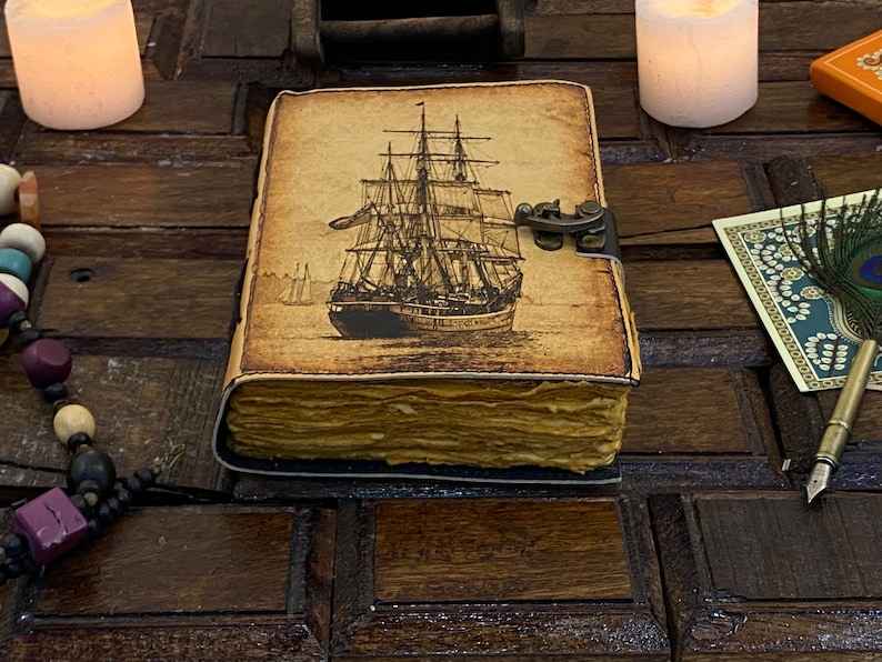 Pode incluir: Um di&aacute;rio de estilo vintage encadernado em couro com uma ilustra&ccedil;&atilde;o detalhada de um navio &agrave; vela na capa. O di&aacute;rio tem um fecho de lat&atilde;o e parece ser antigo e desgastado.