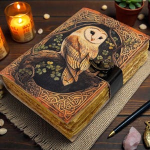 May include: A detailed, leather-bound journal with an owl illustration on the cover. The journal features a clasp closure and aged paper. The image includes candles, a pen, and small potted plants, creating a cozy, artistic aesthetic.
