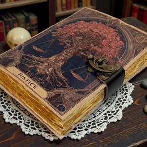 May include: A vintage-style journal with a detailed cover featuring a tree design and scales, with the word "JUSTICE". The journal has aged, yellowed pages and a clasp closure. It rests on a white lace doily.
