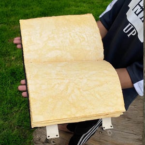 May include: A large, handmade book with a yellowed, textured paper cover. The book is open and the pages are visible. The book is being held by a person's hands.