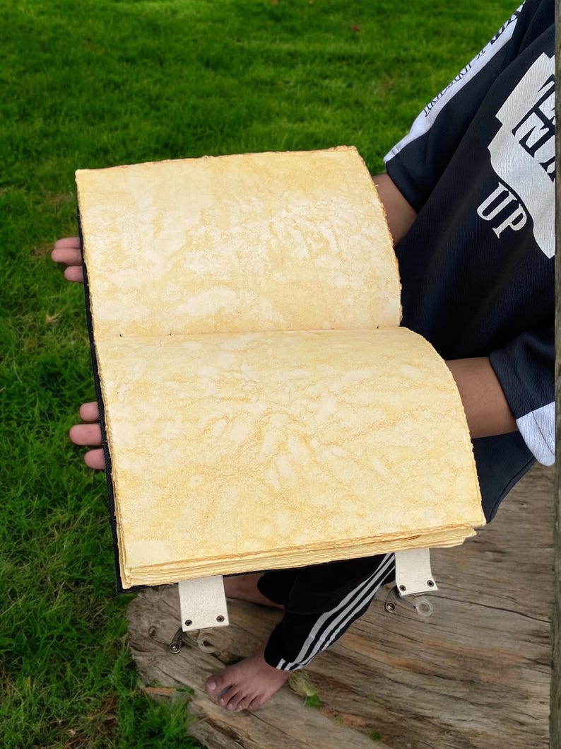 May include: A large, handmade book with a yellowed, textured paper cover. The book is open and the pages are visible. The book is being held by a person's hands.
