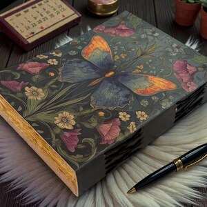 May include: A decorative journal with a butterfly and floral design on the cover. The cover is dark gray with a butterfly in shades of blue and orange. Pink and yellow flowers and green leaves surround the butterfly. A black pen with gold accents rests on a white fur surface.