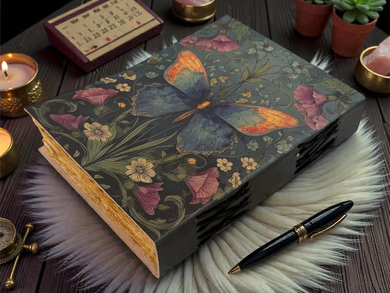 May include: A decorative journal with a butterfly and floral design on the cover. The cover is dark gray with a butterfly in shades of blue and orange. Pink and yellow flowers and green leaves surround the butterfly. A black pen with gold accents rests on a white fur surface.