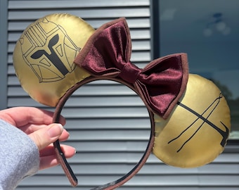 May include: Gold-colored mouse ears headband with a burgundy bow. One ear features a black outline of a helmet, and the other ear has a black graphic. The headband is brown.