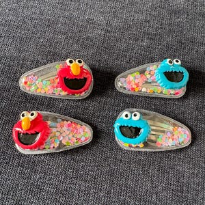 May include: Four clear hair clips with colourful confetti inside. Two clips feature a red character with a large smile, and two feature a blue character with a wide grin. The clips are designed for children.