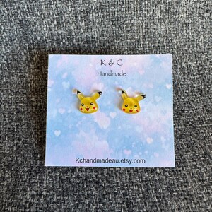 May include: Yellow Pikachu stud earrings with black-tipped ears and red cheeks, presented on a light blue card with a heart pattern. The card features the text "K & C Handmade" and "Kchandmadeau.etsy.com".