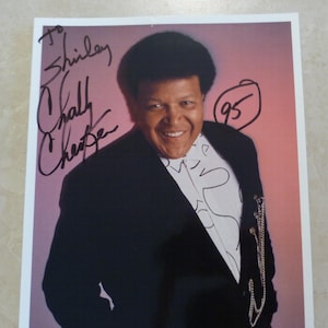 May include: A black and white photograph of Chubby Checker, a singer, wearing a black suit jacket and a white shirt with a patterned design. He is smiling and looking at the camera. The photo is signed "To Shirley Chally Checker 95".