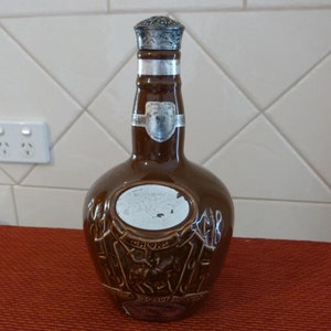 May include: A brown ceramic bottle with a silver top. The bottle has a label that says "Chivas Regal" and a crest with a knight on a horse. The bottle is sitting on a red surface.