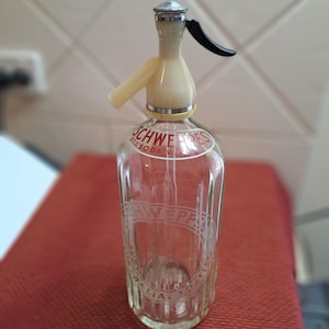 Può includere: Una bottiglia sifone per acqua gassata Schweppes vintage in vetro trasparente. La bottiglia ha una parte superiore color crema con una leva nera e un tappo in metallo argentato. Il logo Schweppes è visibile in rosso e bianco sulla bottiglia.