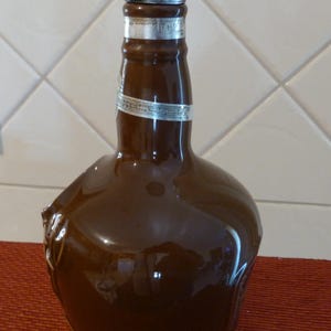 May include: A brown ceramic bottle with a silver top and a silver band around the neck. The bottle has a unique shape with a handle on the side.